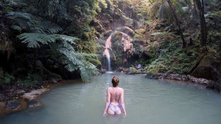 Person wading in a serene forest pond with a waterfall.