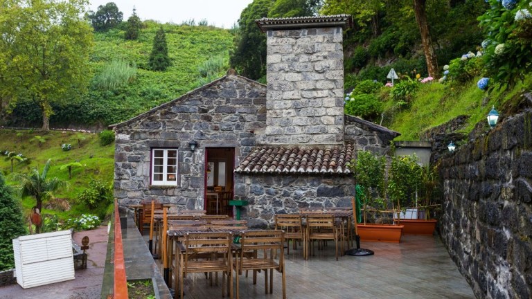 Stone house with terrace and wooden tables, surrounded by greenery.