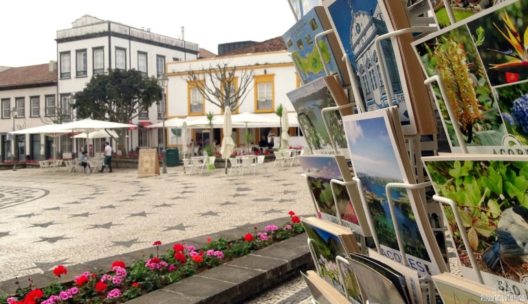 Postcards on display in a plaza with flowerbeds, cafes, and historic buildings in the background.