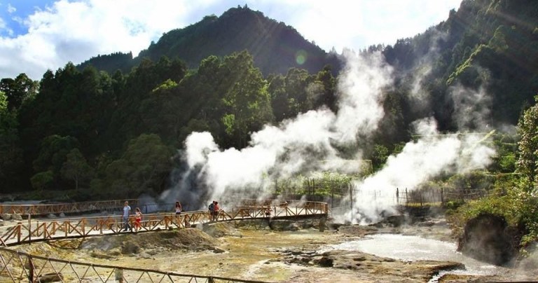 People on a wooden walkway with steam rising near forested hills.