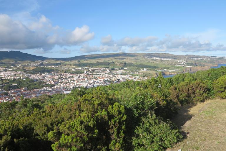 Aerial view of a town with hills, greenery, and scattered clouds in the sky.