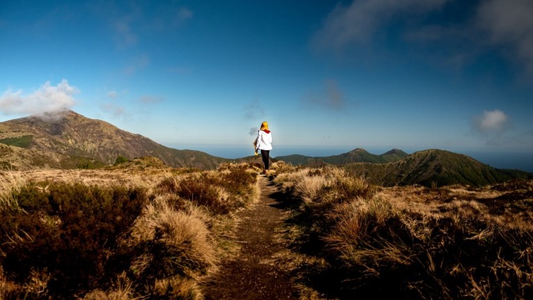 Person walks on mountain trail with a blue sky and distant peaks.