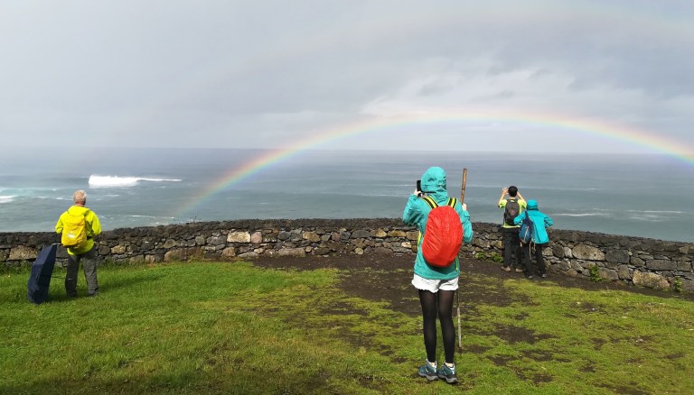 People on grassy area near ocean, photographing a rainbow.