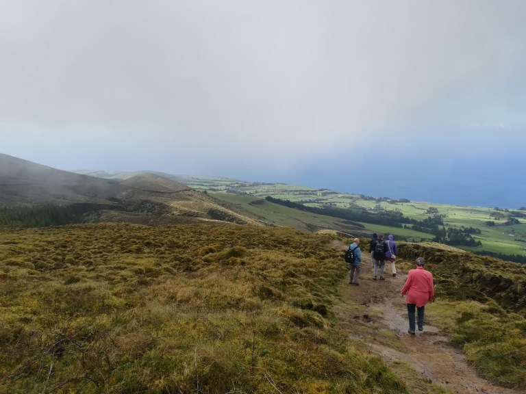 Hikers walking on a grassy path with hills and ocean in the background.