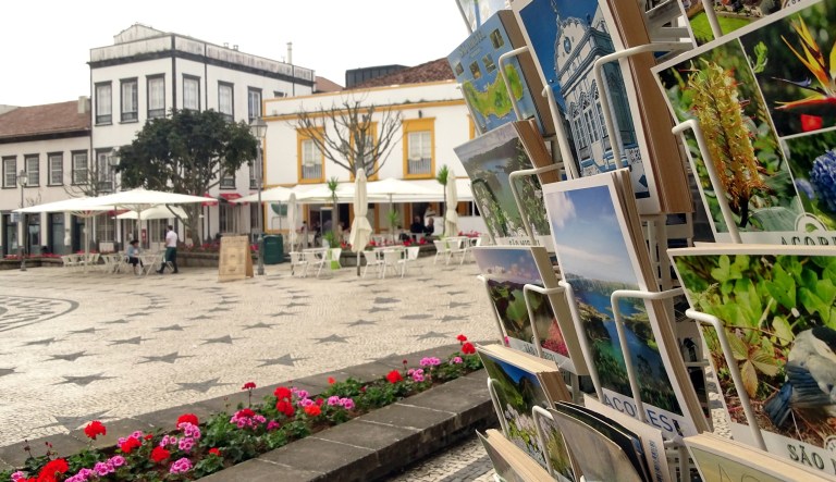 Street scene with postcard rack and outdoor cafe tables on a patterned pavement.