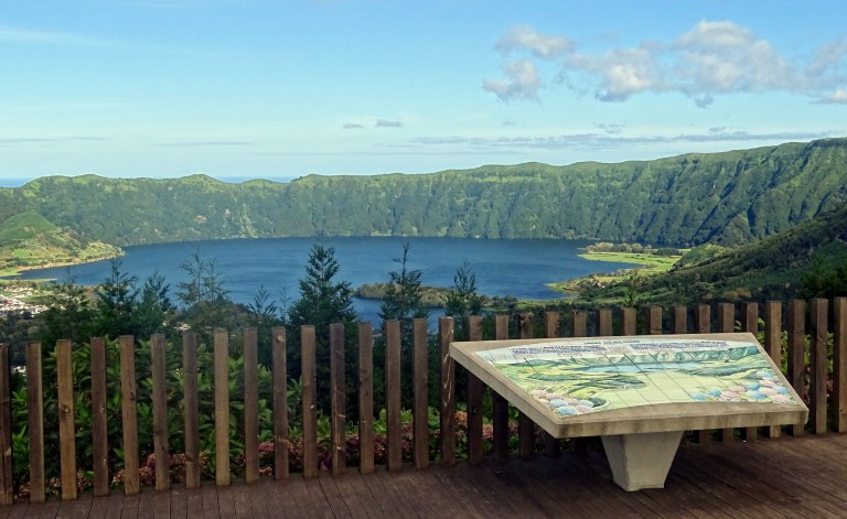 Scenic view of a lake surrounded by lush green hills, with an information board in the foreground.