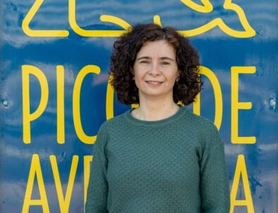 Smiling person with curly hair in front of a blue sign with yellow text.