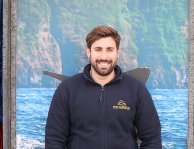 Man in black jacket standing in front of ocean and cliff backdrop.