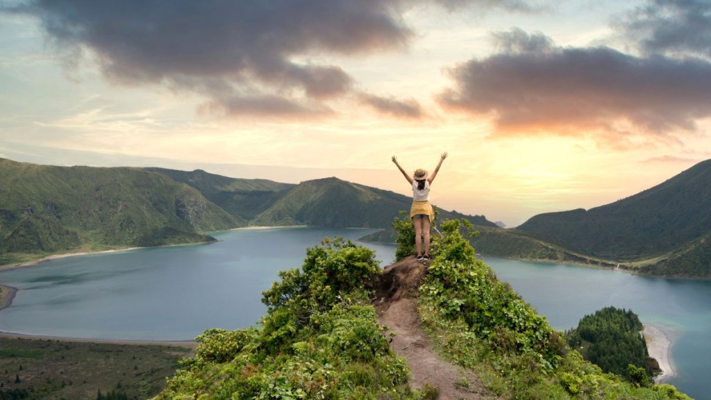 Person with arms raised on a green hill, overlooking a lake and mountains at sunset.