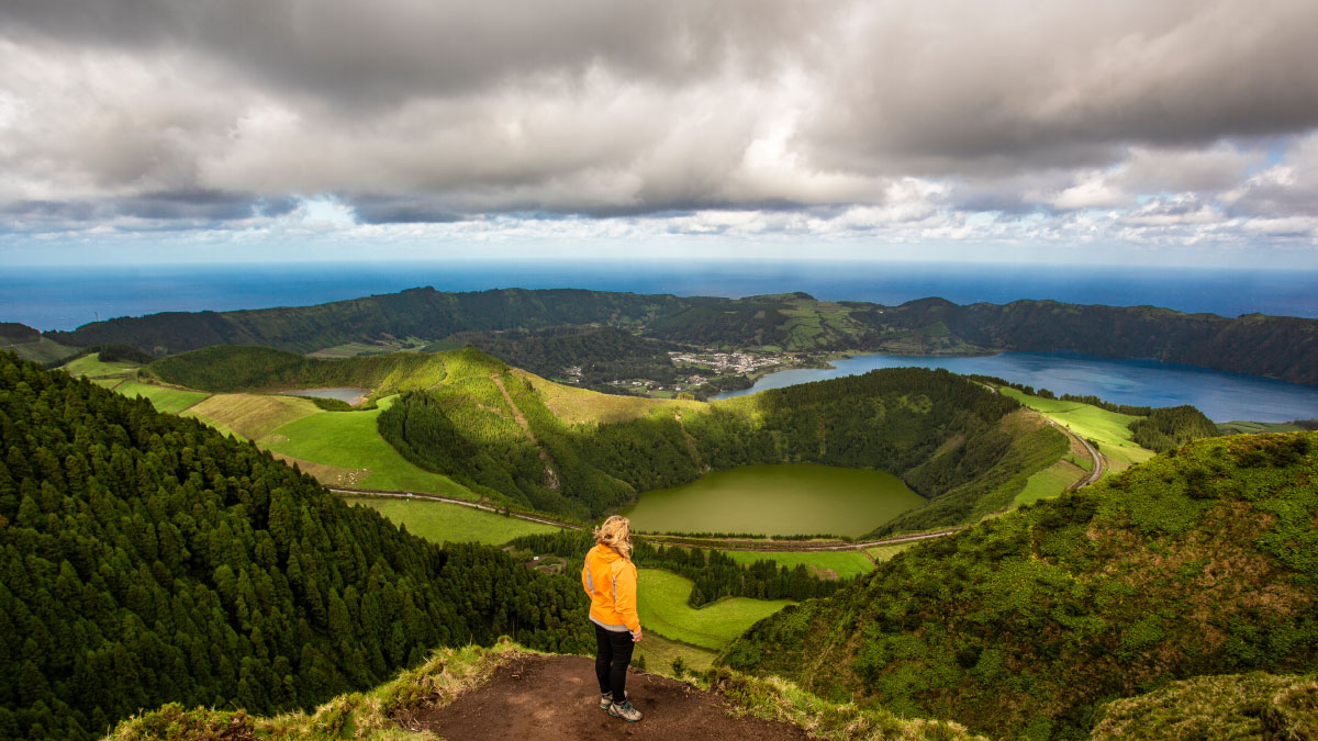 Person in orange jacket standing on hill overlooking lush landscape and lakes under cloudy sky.