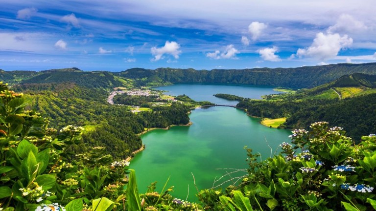 Vibrant lake surrounded by lush green hills under a blue sky with white clouds.