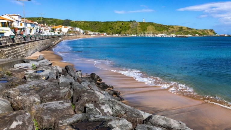 Coastal view with rocky shore, sandy beach, blue sea, and buildings in the distance.