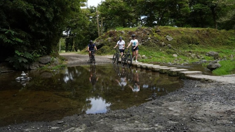 Three cyclists pause at a shallow stream crossing in a wooded area.