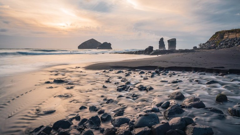 Rocky beach at sunset with distant sea stacks and gentle waves.