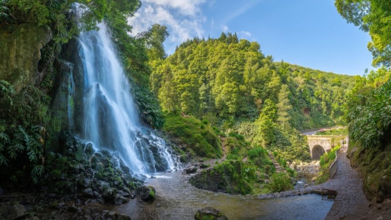 Waterfall flowing into a river with a stone bridge and lush green forest under a blue sky.