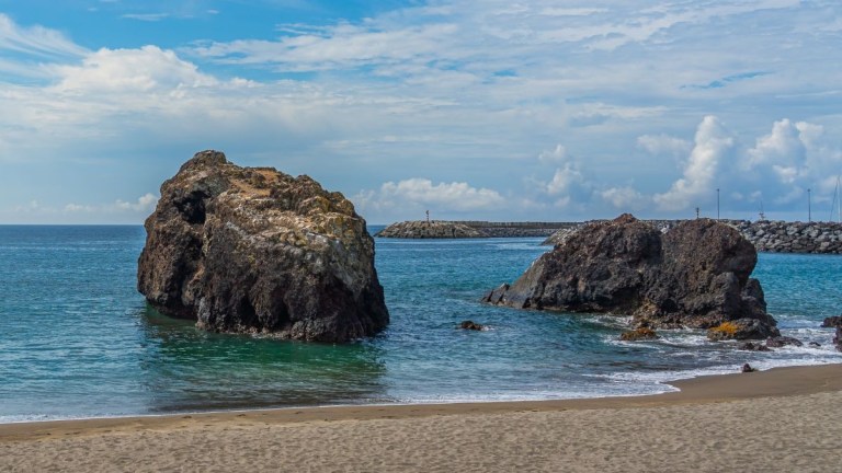 Two large rocks in a calm ocean near a sandy beach.