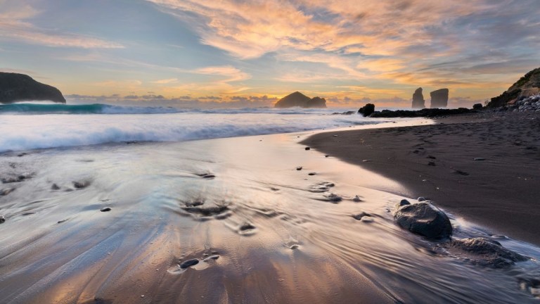 Sunset over a serene beach with gentle waves and distant rock formations.