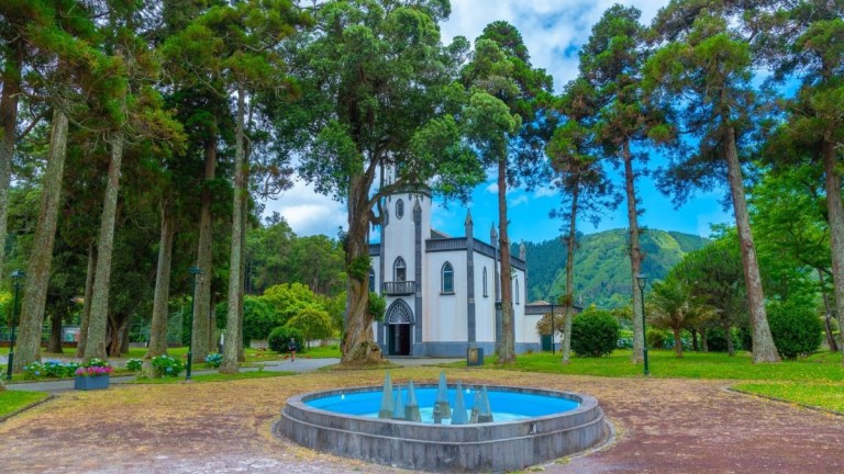White chapel and fountain in a park with tall trees and mountains.