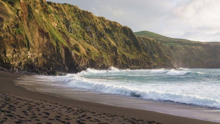 Waves crash on a black sand beach with steep cliffs and lush green hills in the background.