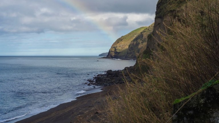 Coastal view with rocky shoreline, hillside, and a rainbow over the ocean.
