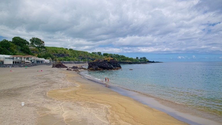 Sandy beach with rocky outcrops, calm ocean, cloudy sky.