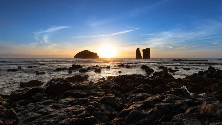 Sunset over ocean with rocky shore and two rock formations in distance.