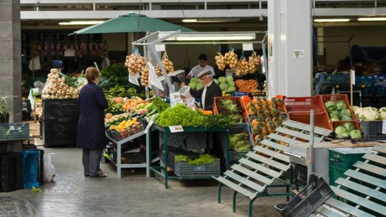 Outdoor market stall with fruits, vegetables, and people shopping.