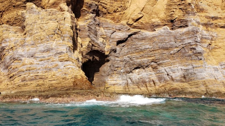 Rocky cliff face with layers and a small cave by the sea.