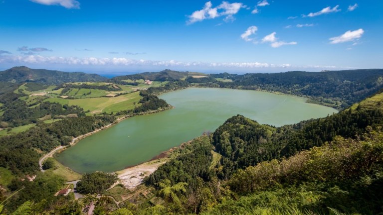 Aerial view of a large green lake surrounded by lush hills and clouds on a sunny day.