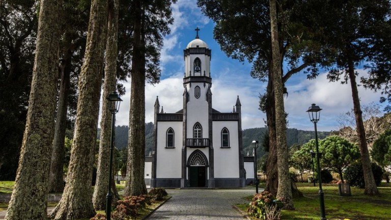 A white church with a bell tower, surrounded by tall trees under a blue sky.