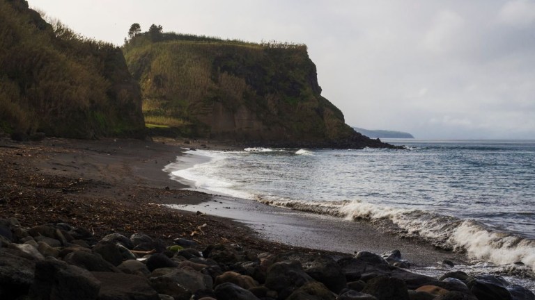 Rocky beach with waves and green cliffs under a cloudy sky.