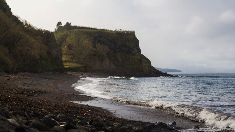 Coastal cliff with rocky beach and waves under a cloudy sky.
