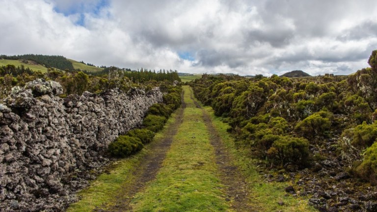 Stone path with mossy walls and cloudy sky in a hilly landscape.