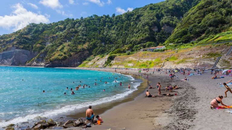 Crowded beach with people swimming and relaxing, lush green hills in the background.