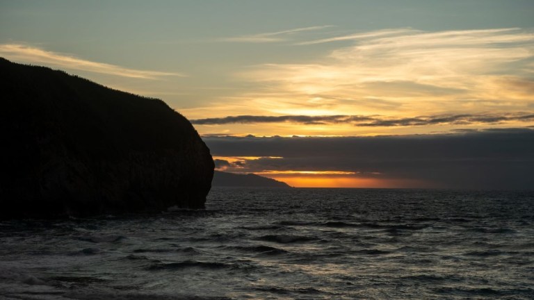 Sunset over ocean, silhouetted cliff in foreground.