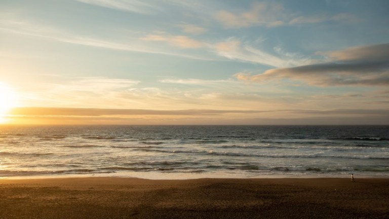 Sun setting over calm ocean waves and sandy beach with cloudy sky.