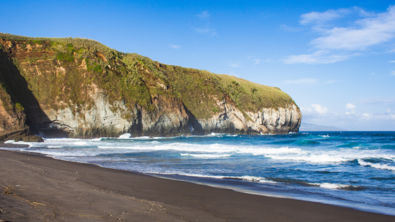 Coastal cliff with grassy top, waves hitting dark sandy beach under a clear blue sky.