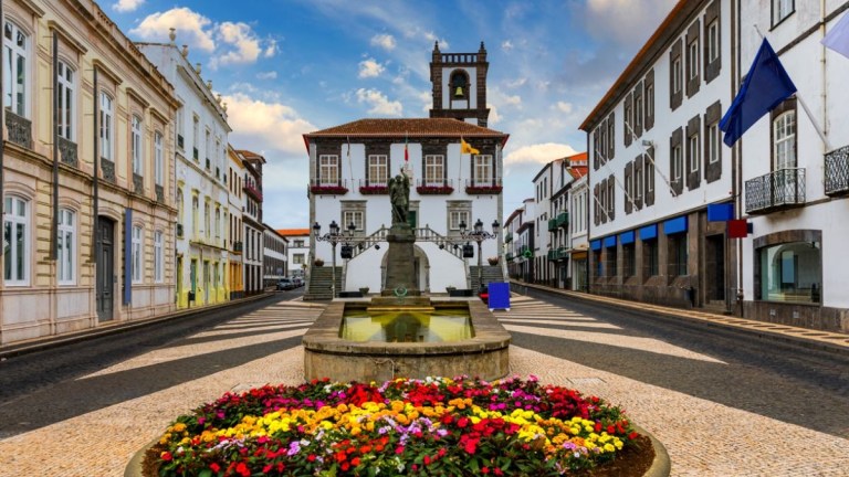 Town square with fountain, colorful flowers, and historic buildings under a blue sky.