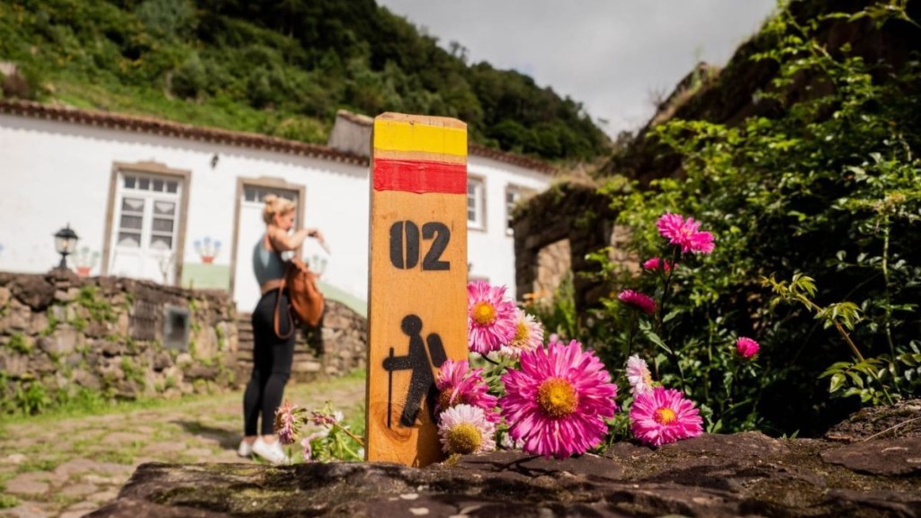 Trail sign with flowers, person and building in background.
