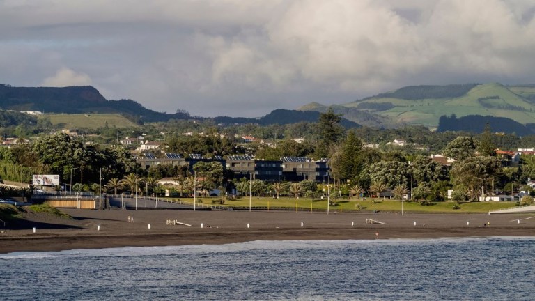 Coastal town with a sandy beach, greenery, and hills in the background under a cloudy sky.