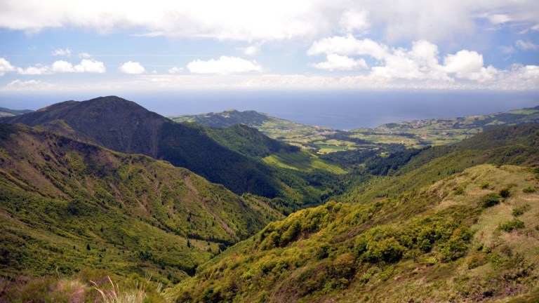 Lush green valley with mountains and ocean under a cloudy sky.