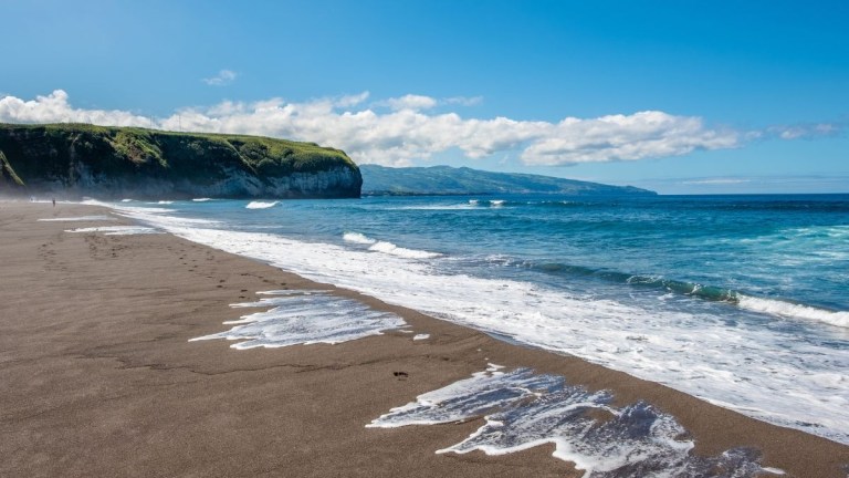 Sandy beach with waves and cliffs under a clear blue sky.