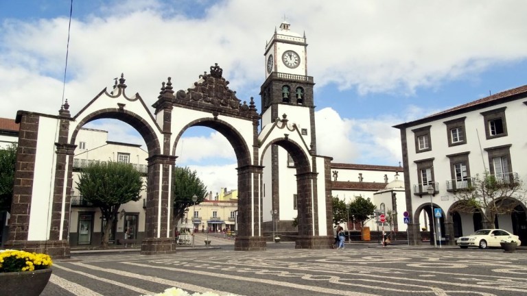 Historic arches and clock tower in a town square with cloudy sky.