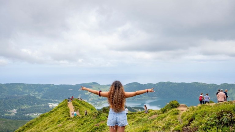 Person with arms outstretched on a hill overlooking lush landscape and lake.