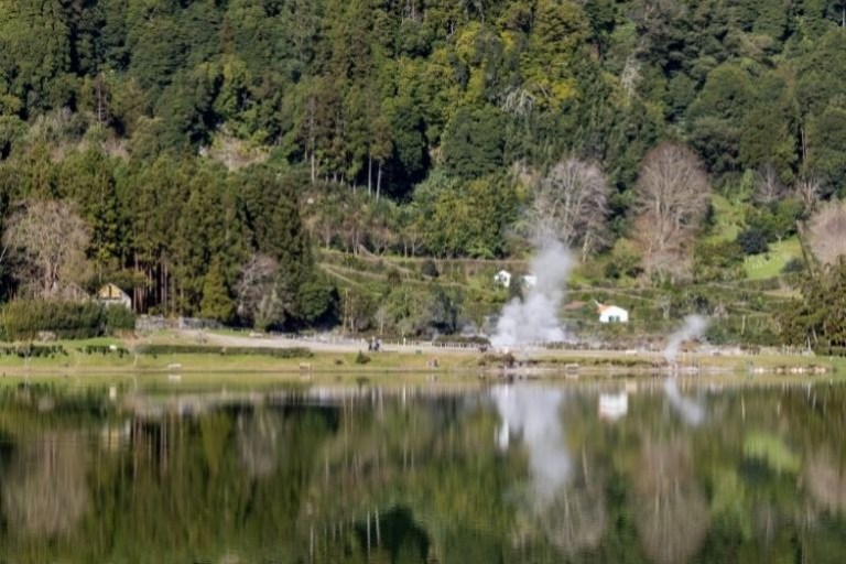 Lakeside view with steam rising and lush forested hills in the background.