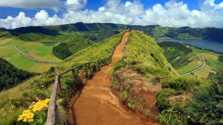 Dirt path with wooden railing on a lush green hillside under a blue sky with clouds.