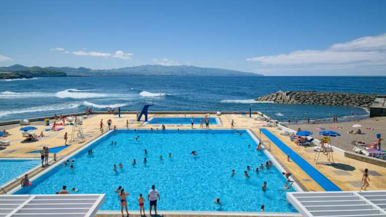 Outdoor pool by the ocean, people swimming, sunny day with umbrellas and distant hills.
