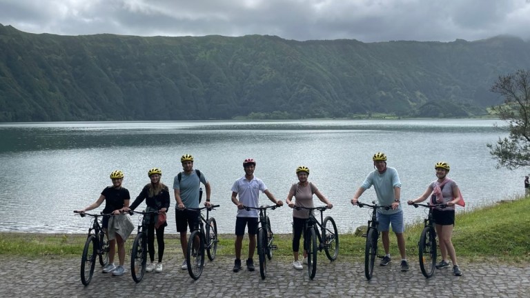Seven people with bikes and helmets by a lake with hills in the background.