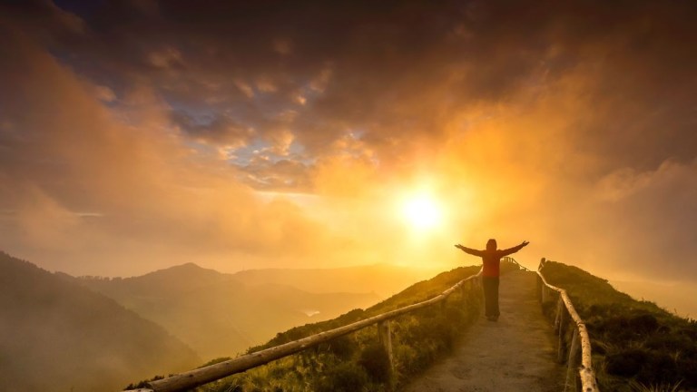 Person with arms raised on a mountain path at sunset.