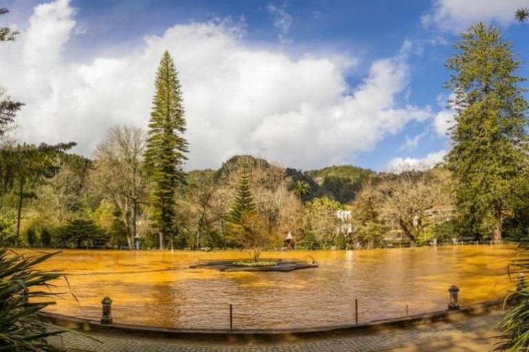 A small pond with orange water surrounded by trees and mountains under a blue sky with clouds.